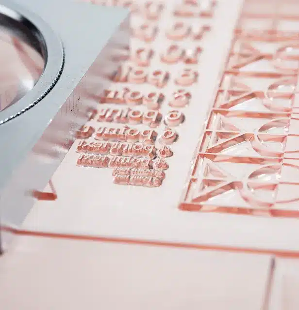 Nahaufnahme einer Klischeeplatte zum Drucken von Texten mit reliefartigen, spiegelverkehrten Buchstaben aus transparentem Kunststoff, daneben eine Lupe.