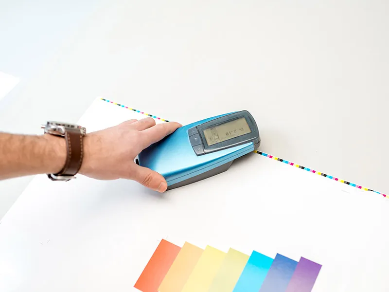 Hand hält ein Farbmessgerät über einem Blatt mit Farbfeldern und Farbkontrollstreifen.