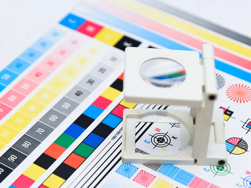 Eine Lupe liegt auf einem Farbdruck-Bogen mit diversen Farbfeldern, Testmustern und Kontrollstreifen für den Offsetdruck.