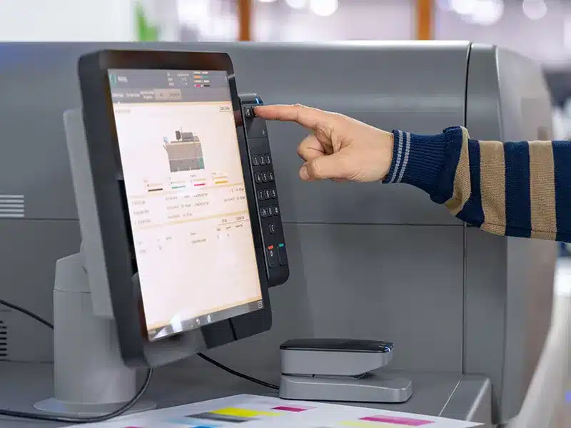 Person tippt mit dem Finger auf einen Touchscreen-Bedienpanel einer modernen Druckmaschine, daneben sind Papierproben und Farbfelder zu sehen.