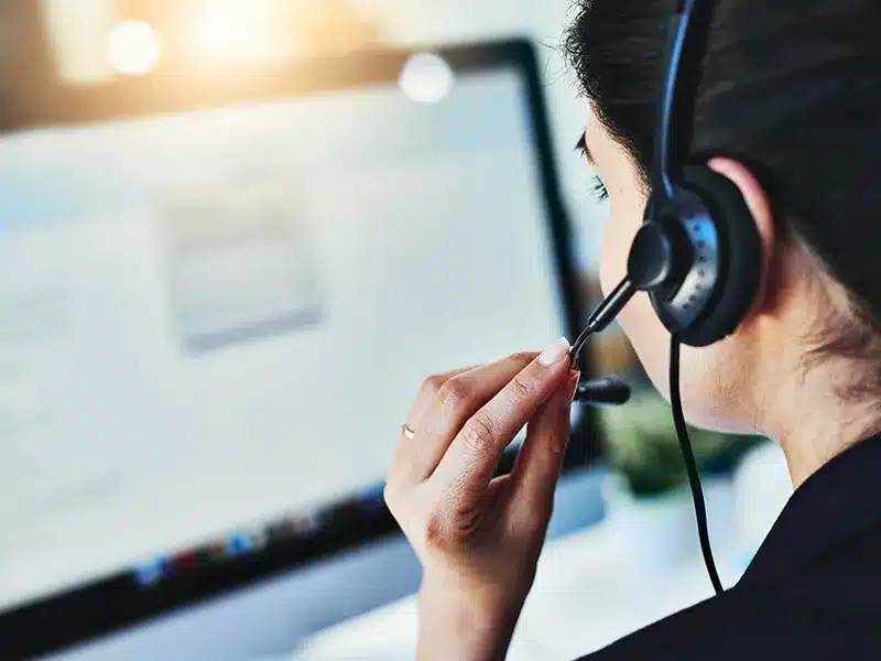 Frau mit Headset sitzt vor einem Computerbildschirm und führt vermutlich ein Telefongespräch oder Kundensupport durch.