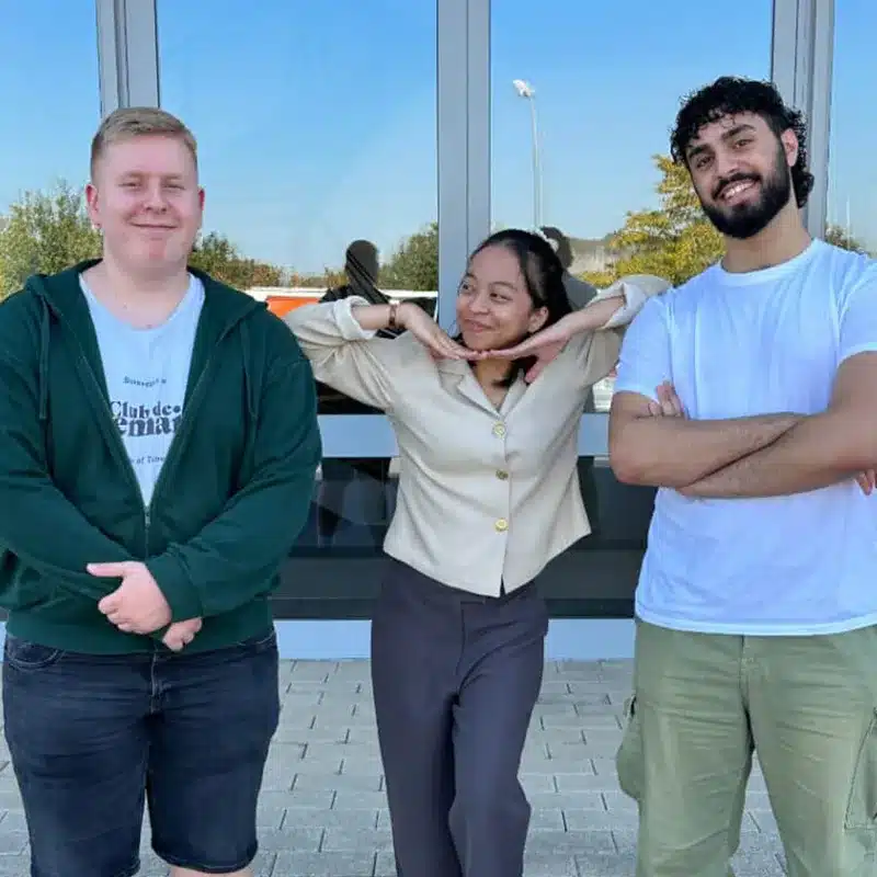 Drei junge Erwachsene posieren lächelnd vor einem großen Fenster; die Person in der Mitte steht mit Händen unter dem Kinn und einem witzigen Gesichtsausdruck, während die anderen entspannt daneben stehen.