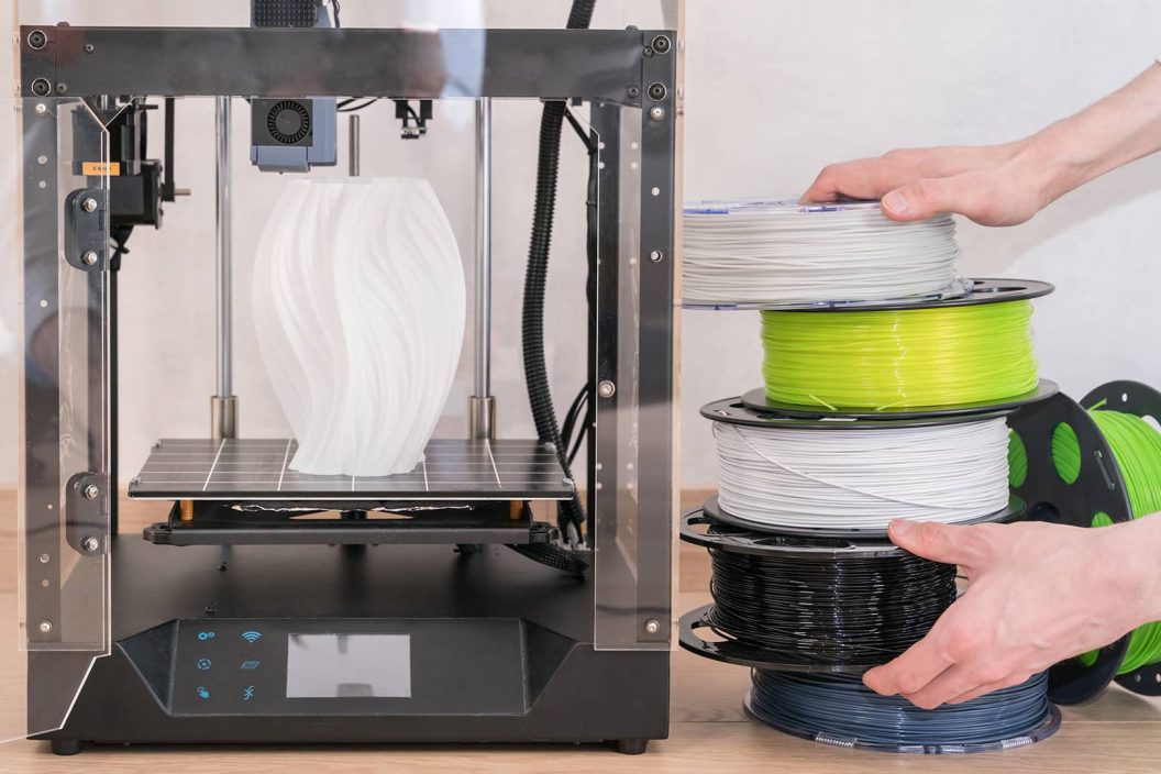 3D-Drucker mit einem weißen, gewellten Objekt auf der Druckplatte neben mehreren Rollen Filament in verschiedenen Farben, die von Händen gestapelt werden.